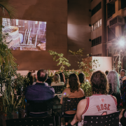 A imagem retrata uma sessão de cinema ao ar livre em um espaço urbano durante a noite. No fundo, há prédios altos com fachadas iluminadas por luzes artificiais, além de uma estrutura de madeira com plantas trepadeiras e iluminação destacada. Algumas plantas e arbustos em vasos estão espalhados pelo local, trazendo um toque de natureza ao ambiente. No centro da imagem, uma projeção é exibida em uma parede clara, mostrando uma cena com utensílios de cozinha e legendas em português. As legendas dizem: "Eu vou olhar a armadilha e aproveito para trazer lenha."
Em primeiro plano, várias pessoas estão sentadas em cadeiras pretas de plástico, voltadas para a projeção.