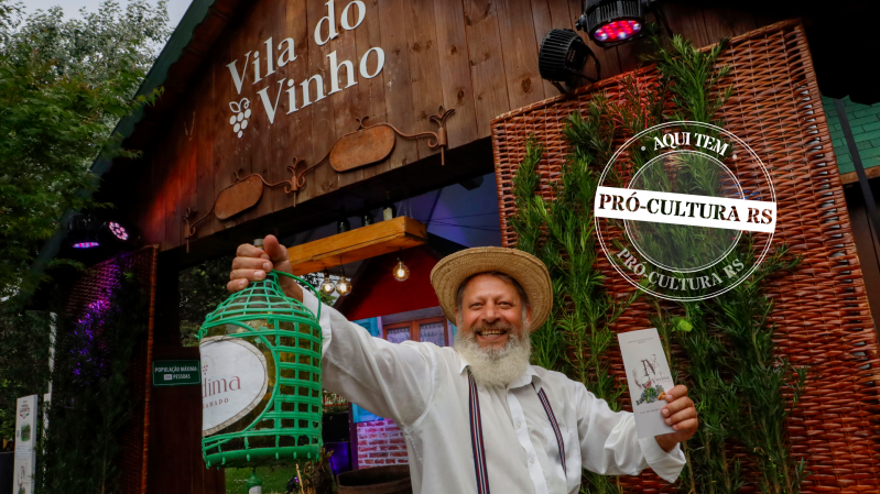 Um homem com trajes italianos segura um garrafão de vinho em frente a uma casa de madeira na qual está escrito "Vila do Vinho"