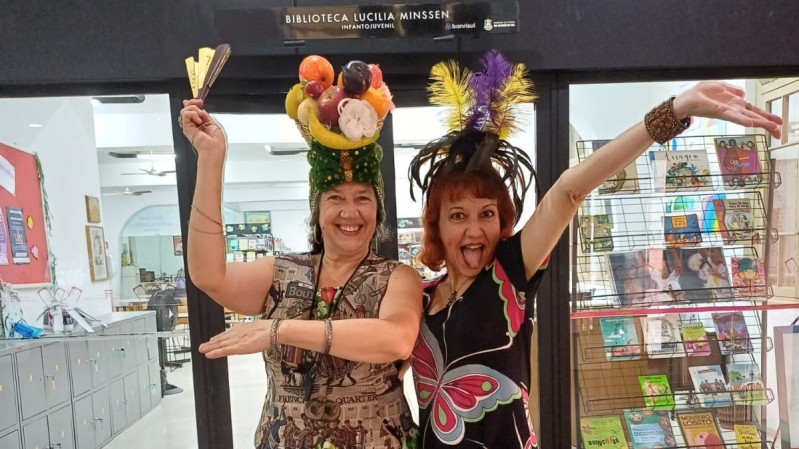 Duas mulhres estão em pé e lado a lado, com adereços carnavalescos, em frente à Biblioteca Lucília Minssen