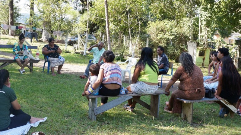 Num gramado cercado de árvores, sentados em bancos de madeira colocados em círculo, homens, mulheres e crianças escutam orador de camisa branca ao centro da imagem