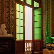 Interior de uma sala decorada com estilo clássico e elementos ornamentais. Ao centro, uma porta de madeira com vidros coloridos, predominantemente verdes e amarelos, está aberta, revelando parcialmente uma varanda com balaústres de pedra. À esquerda, há uma escultura de uma esfinge branca sobre um pedestal escuro, com inscrições em português. As paredes são decoradas com padrões detalhados em cores vibrantes, como dourado, verde e vermelho. À direita, parte de um sofá antigo com encosto entalhado e estofado claro é visível.