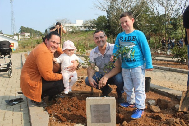 Em uma área aberta, a imagem mostra uma família em um canteiro diante de uma muda de árvore recém plantada.