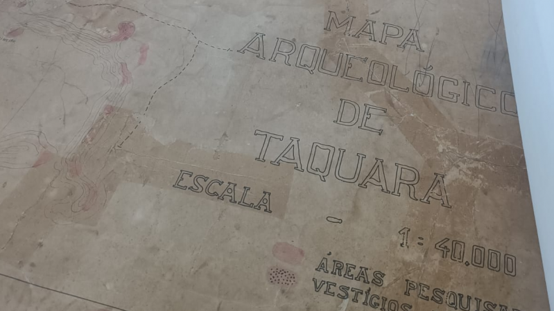 Fotografia de um mapa arqueológico antigo, com papel envelhecido. No canto direito, lê-se o título “Mapa Arqueológico de Taquara” com a indicação de escala “1:40.000”. Abaixo, está escrito “Áreas pesquisadas – vestígios indígenas”. O mapa mostra contornos topográficos em linhas pretas e áreas destacadas em vermelho, possivelmente indicando locais de interesse arqueológico.