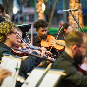 Fotografia colorida de uma apresentação ao ar livre da orquestra OSPA Jovem. A imagem mostra vários músicos concentrados tocando instrumentos de corda, como violinos e violas. Todos estão vestidos com roupas escuras e partituras estão apoiadas à frente deles. A expressão dos músicos indica concentração e envolvimento com a performance.
