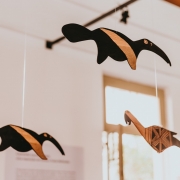 Três esculturas de madeira em forma de tamanduás estão suspensas por fios no teto de uma sala expositiva. As peças têm desenhos geométricos pintados em preto e marrom, criando um contraste elegante. Ao fundo, a luz natural entra pelas janelas, iluminando suavemente o ambiente.