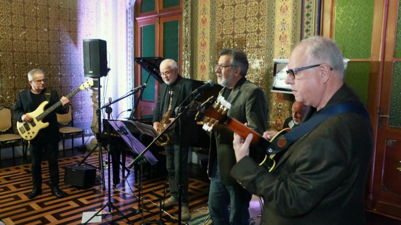 Cinco músicos se apresentam em uma sala com decoração clássica. À esquerda, um homem toca contrabaixo elétrico; ao centro, outro músico toca saxofone e há um vocalista ao lado dele. À direita, um guitarrista toca violão elétrico. Todos usam roupas escuras. O ambiente tem piso de madeira com padrão geométrico e paredes ricamente decoradas.