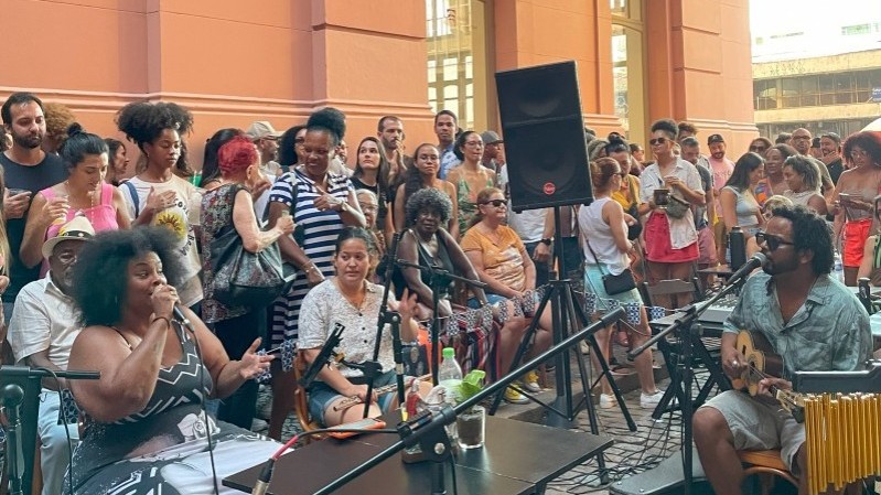 Uma roda de samba acontece ao ar livre diante de um prédio histórico de cor rosa, com colunas e sacadas ornamentadas. Na cena, uma cantora negra com vestido preto e branco canta ao microfone à esquerda, enquanto um músico toca cavaquinho à direita. Há instrumentos de percussão em primeiro plano. Um grande grupo de pessoas assiste à apresentação, reunido em pé e sentado, demonstrando interesse e alegria. O ambiente é descontraído e festivo, com clima de fim de tarde. A estrutura do prédio e a disposição do público sugerem que o evento ocorre em um espaço cultural urbano.