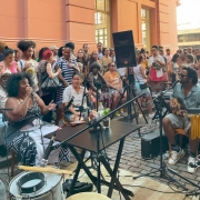 Uma roda de samba acontece ao ar livre diante de um prédio histórico de cor rosa, com colunas e sacadas ornamentadas. Na cena, uma cantora negra com vestido preto e branco canta ao microfone à esquerda, enquanto um músico toca cavaquinho à direita. Há instrumentos de percussão em primeiro plano. Um grande grupo de pessoas assiste à apresentação, reunido em pé e sentado, demonstrando interesse e alegria. O ambiente é descontraído e festivo, com clima de fim de tarde. A estrutura do prédio e a disposição do público sugerem que o evento ocorre em um espaço cultural urbano.