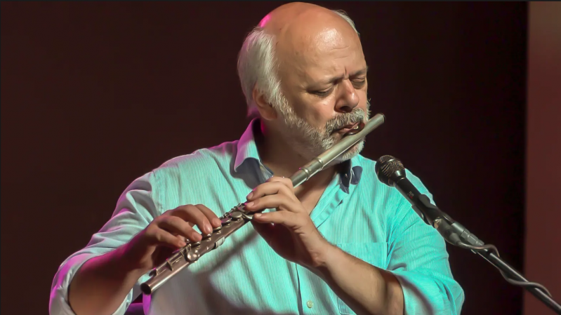 A imagem mostra o flautista, saxofonista, compositor e arranjador Pedrinho Figueiredo: ele tem pele clara, cabelos, barba e bigode brancos. O homem está tocando uma flauta transversal de metal prateado e parece estar profundamente concentrado na música. Veste uma camisa azul-clara com colarinho aberto e está diante de um microfone. O fundo é escuro e desfocado, destacando o músico com iluminação em tons quentes e suaves, sugerindo um ambiente de apresentação ao vivo, como um concerto ou show intimista.