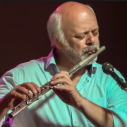 A imagem mostra o flautista, saxofonista, compositor e arranjador Pedrinho Figueiredo: ele tem pele clara, cabelos, barba e bigode brancos. O homem está tocando uma flauta transversal de metal prateado e parece estar profundamente concentrado na música. Veste uma camisa azul-clara com colarinho aberto e está diante de um microfone. O fundo é escuro e desfocado, destacando o músico com iluminação em tons quentes e suaves, sugerindo um ambiente de apresentação ao vivo, como um concerto ou show intimista.