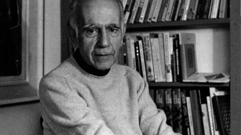 Homem idoso de pele clara, cabelos curtos e grisalhos, veste um suéter claro e está sentado em um ambiente interno. Ele olha diretamente para a câmera com uma expressão séria e tranquila. Ao fundo, há uma estante de madeira com diversos livros organizados, sugerindo um ambiente de estudo ou escritório. A imagem está em preto e branco.