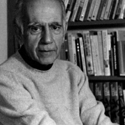 Homem idoso de pele clara, cabelos curtos e grisalhos, veste um suéter claro e está sentado em um ambiente interno. Ele olha diretamente para a câmera com uma expressão séria e tranquila. Ao fundo, há uma estante de madeira com diversos livros organizados, sugerindo um ambiente de estudo ou escritório. A imagem está em preto e branco.