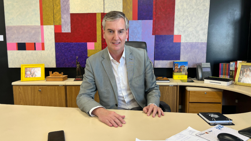 A imagem mostra o novo secretário de Estado da Cultura, Eduardo Loureiro. Ele está vestindo terno cinza claro e camisa branca, sentado atrás de uma mesa em um escritório. Ele está sorrindo levemente e olhando diretamente para a câmera. Sobre a mesa há papéis, um celular, um mouse, uma caneta e um caderno preto. Atrás dele, há uma parede com uma grande obra de arte geométrica e colorida, além de objetos decorativos, porta-retratos com fotos de família, livros, uma escultura e um telefone fixo. O ambiente transmite um ar profissional e organizado, com um toque pessoal.
