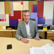 A imagem mostra o novo secretário de Estado da Cultura, Eduardo Loureiro. Ele está vestindo terno cinza claro e camisa branca, sentado atrás de uma mesa em um escritório. Ele está sorrindo levemente e olhando diretamente para a câmera. Sobre a mesa há papéis, um celular, um mouse, uma caneta e um caderno preto. Atrás dele, há uma parede com uma grande obra de arte geométrica e colorida, além de objetos decorativos, porta-retratos com fotos de família, livros, uma escultura e um telefone fixo. O ambiente transmite um ar profissional e organizado, com um toque pessoal.