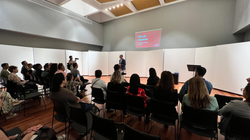 A imagem mostra o secretário de Estado da Cultura, Eduardo Loureiro, de pé, no centro da Sala de Recitais da Casa da Ospa. No fundo, há um telão com a apresentação projetada, exibindo o título “Fala de Abertura” e o nome do palestrante, “Secretário Eduardo Loureiro”. O público está sentado em cadeiras organizadas em fileiras, prestando atenção. À esquerda, um fotógrafo registra o momento. O ambiente possui paredes brancas, piso de madeira e iluminação embutida no teto. A atmosfera é profissional e voltada para uma apresentação formal ou institucional.