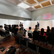 A imagem mostra o secretário de Estado da Cultura, Eduardo Loureiro, de pé, no centro da  Sala de Recitais da Casa da Ospa. No fundo, há um telão com a apresentação projetada, exibindo o título “Fala de Abertura” e o nome do palestrante, “Secretário Eduardo Loureiro”. O público está sentado em cadeiras organizadas em fileiras, prestando atenção. À esquerda, um fotógrafo registra o momento. O ambiente possui paredes brancas, piso de madeira e iluminação embutida no teto. A atmosfera é profissional e voltada para uma apresentação formal ou institucional.