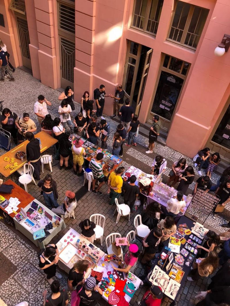 A imagem mostra uma feira ao ar livre em um espaço urbano entre prédios de arquitetura clássica, com paredes rosa claro e janelas altas. As pessoas circulam por corredores formados por diversas mesas retangulares onde expositores vendem itens artesanais e criativos, como bijuterias, ilustrações, acessórios coloridos e objetos decorativos. O ambiente é descontraído e diverso, com um público majoritariamente jovem, vestindo roupas alternativas. Algumas pessoas estão sentadas, outras observam os produtos ou conversam.