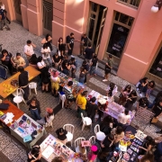 A imagem mostra uma feira ao ar livre em um espaço urbano entre prédios de arquitetura clássica, com paredes rosa claro e janelas altas. As pessoas circulam por corredores formados por diversas mesas retangulares onde expositores vendem itens artesanais e criativos, como bijuterias, ilustrações, acessórios coloridos e objetos decorativos. O ambiente é descontraído e diverso, com um público majoritariamente jovem, vestindo roupas alternativas. Algumas pessoas estão sentadas, outras observam os produtos ou conversam.
