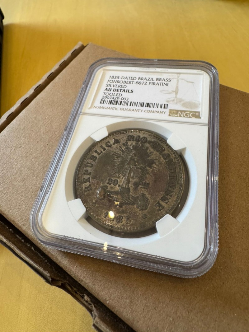 Close-up de uma moeda antiga brasileira encapsulada e certificada pela Numismatic Guaranty Company (NGC). O rótulo diz:
“1835-Dated Brazil Brass
Fonrobert-8872 Piratini
Silvered
AU Details
Tooled
20907429-003”.
A moeda tem a inscrição “REPUBLICA RIOGRANDENSE” com uma balança, raios de sol e a data de 1835 em destaque. A peça está acondicionada em um estojo plástico sobre uma superfície de papelão.