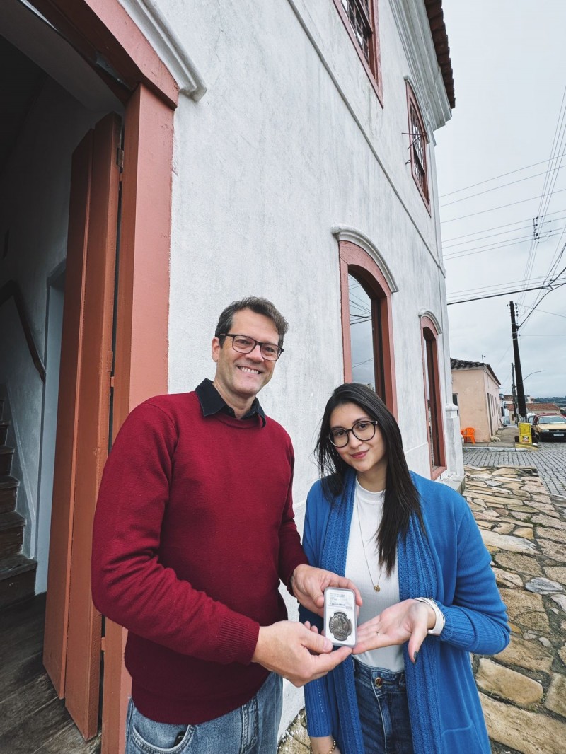 Duas pessoas estão em pé na frente de um prédio histórico com fachada branca e janelas de madeira vermelha. À esquerda, um homem de óculos, vestindo suéter vermelho e jeans, sorri enquanto segura uma cápsula transparente com uma moeda antiga. À direita, uma mulher de cabelos longos e escuros, também de óculos, usa um casaco azul sobre uma blusa clara e segura a mesma cápsula com uma das mãos estendida. Ambos posam felizes para a foto.