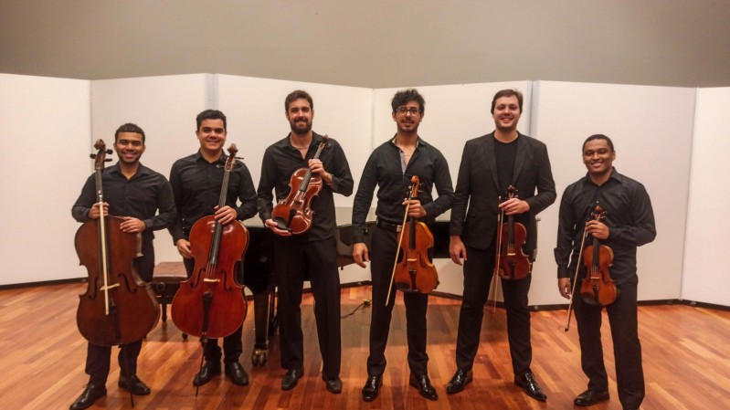 Seis músicos estão em pé lado a lado, posando para uma foto em um ambiente interno com piso de madeira e painéis brancos ao fundo. Todos vestem roupas formais escuras e seguram instrumentos de corda. Da esquerda para a direita: O primeiro e o segundo músicos seguram violoncelos, o terceiro segura uma viola, o quarto, quinto e sexto seguram violinos.
Ao fundo, há um piano de cauda parcialmente visível. 