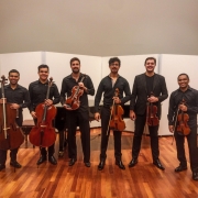 Seis músicos estão em pé lado a lado, posando para uma foto em um ambiente interno com piso de madeira e painéis brancos ao fundo. Todos vestem roupas formais escuras e seguram instrumentos de corda. Da esquerda para a direita: O primeiro e o segundo músicos seguram violoncelos, o terceiro segura uma viola, o quarto, quinto e sexto seguram violinos.
Ao fundo, há um piano de cauda parcialmente visível. 