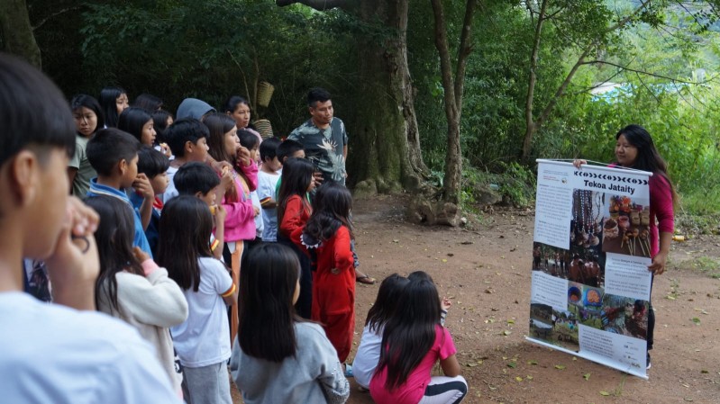 Uma grande multidão de crianças está ouvindo uma mulher que apresenta um painel.
A composição da cena mostra um grupo numeroso de crianças e adolescentes, em grande parte posicionados de frente para uma mulher, que está segurando um painel com informações. A mulher está no centro da imagem, segurando o painel e olhando para as crianças. A posição das crianças sugere escuta atenta e interesse no conteúdo apresentado. A atmosfera parece informal e educacional, com foco em uma troca de conhecimentos. A imagem mostra uma visão de frente para o grupo de crianças, em um local ao ar livre, com árvores ao fundo.
O local parece ser um terreno aberto com árvores e vegetação ao redor. A iluminação é natural, provinda do sol. O tom geral da imagem é de interação positiva entre adultos e crianças e transmite uma atmosfera educativa.