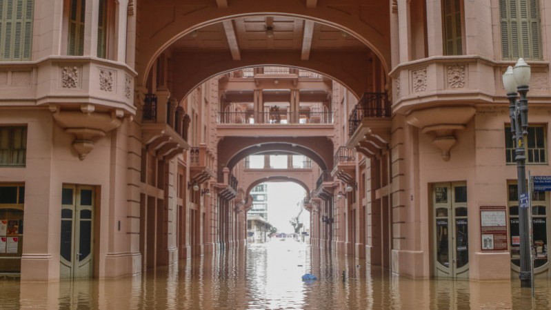 Foto de uma rua alagada entre prédios históricos em estilo clássico com arcadas. A água cobre completamente o chão, refletindo as fachadas cor-de-rosa dos edifícios.