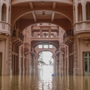 Foto de uma rua alagada entre prédios históricos em estilo clássico com arcadas. A água cobre completamente o chão, refletindo as fachadas cor-de-rosa dos edifícios.