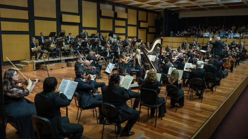Fotografia da Orquestra Sinfônica de Porto Alegre (Ospa), em apresentação em uma sala de concertos no Complexo Cultural da Orquestra, em Porto Alegre. Os músicos estão elegantemente vestidos com trajes formais, predominantemente em preto. Estão distribuídos por naipes: à frente, os instrumentos de cordas (violinos, violas, violoncelos e contrabaixos); ao centro e à direita, instrumentos de sopro e percussão; e ao fundo, em um nível elevado, estão os metais e a percussão. Em destaque próximo ao centro da imagem há uma harpa. O maestro está de pé, à direita, sobre um pequeno pódio, regendo a orquestra. Ao fundo, o público acompanha a apresentação de um camarote elevado. O ambiente tem piso de madeira, paredes com tratamento acústico e um teto com painéis geométricos.