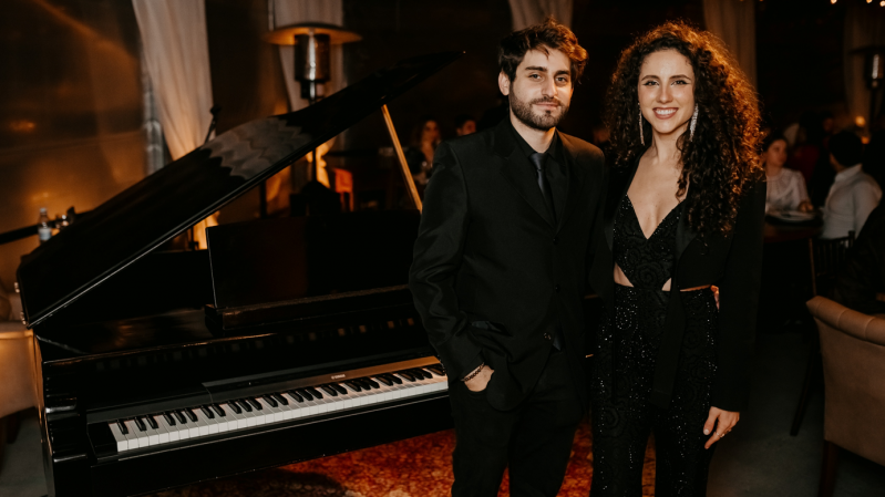 Na imagem, vemos os músicos Cristian Sperandir e Laura Dalmás posando ao lado de um piano preto, em um ambiente elegante e com iluminação quente e suave, sugerindo uma apresentação musical intimista.
O homem está vestido com um terno preto, camisa preta e gravata preta, com as mãos nos bolsos, enquanto a mulher usa um macacão preto brilhante com detalhes em recortes e um blazer por cima. Ela tem cabelos longos e cacheados, usa brincos e está sorrindo. Ao fundo, é possível ver outras pessoas sentadas, cortinas brancas e luzes de festa penduradas, contribuindo para o clima acolhedor e refinado da ocasião.