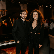 Na imagem, vemos os músicos Cristian Sperandir e Laura Dalmás posando ao lado de um piano  preto, em um ambiente elegante e com iluminação quente e suave, sugerindo uma apresentação musical intimista.

O homem está vestido com um terno preto, camisa preta e gravata preta, com as mãos nos bolsos, enquanto a mulher usa um macacão preto brilhante com detalhes em recortes e um blazer por cima. Ela tem cabelos longos e cacheados, usa brincos e está sorrindo. Ao fundo, é possível ver outras pessoas sentadas, cortinas brancas e luzes de festa penduradas, contribuindo para o clima acolhedor e refinado da ocasião.
