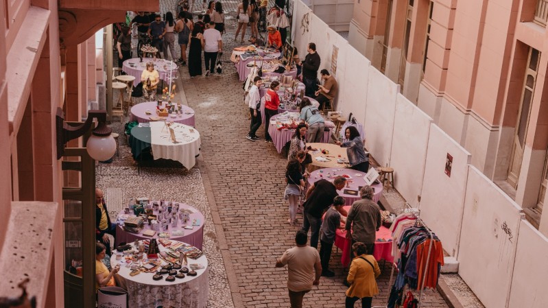Vista aérea de uma feira ao ar livre em uma rua estreita de paralelepípedos, cercada por prédios de fachada rosada. Diversas mesas redondas, cobertas com toalhas coloridas (principalmente em tons de roxo e branco), estão dispostas ao longo da calçada e da rua, exibindo artesanatos, livros, roupas e outros produtos. Várias pessoas circulam entre as barracas, conversando e observando os itens à venda.