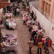 Vista aérea de uma feira ao ar livre em uma rua estreita de paralelepípedos, cercada por prédios de fachada rosada. Diversas mesas redondas, cobertas com toalhas coloridas (principalmente em tons de roxo e branco), estão dispostas ao longo da calçada e da rua, exibindo artesanatos, livros, roupas e outros produtos. Várias pessoas circulam entre as barracas, conversando e observando os itens à venda.