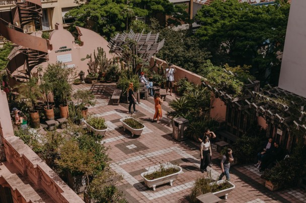 Vista superior do Jardim Lutzenberger, um jardim urbano localizado no terraço de um prédio. O espaço é decorado com plantas diversas em vasos e em banheiras brancas reutilizadas como jardineiras. Há caminhos de ladrilhos em tons de rosa, branco e marrom formando desenhos geométricos. Diversas pessoas caminham, conversam ou descansam em bancos de madeira espalhados pelo local. À esquerda, uma escadaria em espiral dá acesso ao jardim. Ao fundo, árvores e a vegetação urbana completam o cenário.