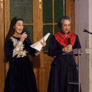 Duas mulheres em pé, lado a lado, falando ao microfone em um ambiente fechado, provavelmente durante um evento formal ou cerimônia. A mulher à esquerda tem cabelos longos e lisos, veste um vestido azul-escuro de veludo com bordados dourados e segura folhas de papel em uma das mãos enquanto fala de forma expressiva ao microfone. A mulher à direita tem cabelos grisalhos presos, usa óculos e está vestida com um vestido azul-escuro com detalhes em vermelho no colo, além de segurar um microfone