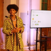 um homem em pé diante de um púlpito transparente e próximo a uma tela digital. Ele veste um conjunto colorido e estampado com padrões geométricos em tons de amarelo, roxo e preto, com o peito parcialmente exposto e adornado com um colar espesso. O homem tem cabelo crespo, volumoso, e está sorrindo levemente, com as mãos entrelaçadas em frente ao corpo, em um gesto descontraído. Ao fundo, há portas de madeira e uma parede com colunas brancas iluminadas. Na tela ao lado dele, aparecem logotipos de patrocinadores e apoiadores institucionais, incluindo o Ministério da Cultura do Brasil e a Shell.