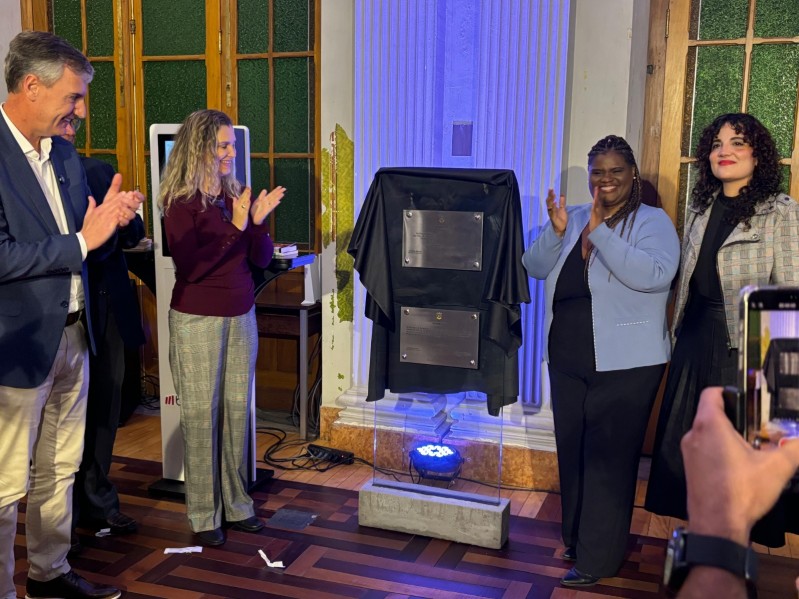 um grupo de cinco pessoas em um ambiente interno, elegantemente decorado, aplaudindo diante de uma estrutura com duas placas metálicas parcialmente cobertas por um tecido preto. O momento parece marcar uma cerimônia de inauguração ou homenagem. No centro da imagem, uma mulher de blazer azul claro e blusa preta sorri enquanto aplaude, ladeada por outras pessoas bem-vestidas — duas mulheres à direita e dois homens à esquerda, um deles usando blazer azul e calça clara. Ao fundo, colunas brancas e portas de madeira com vidros verdes.