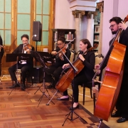  um grupo de músicos clássicos se apresentando em um ambiente interno elegante, possivelmente uma biblioteca ou salão histórico. São cinco músicos sentados e um em pé, formando um pequeno conjunto de câmara. Eles tocam instrumentos de cordas: dois violinos, uma viola, um violoncelo e um contrabaixo. Todos estão vestidos de forma formal, com roupas pretas. Ao fundo, é possível ver estantes com livros, colunas decoradas e janelas de madeira com vidro fosco. 
