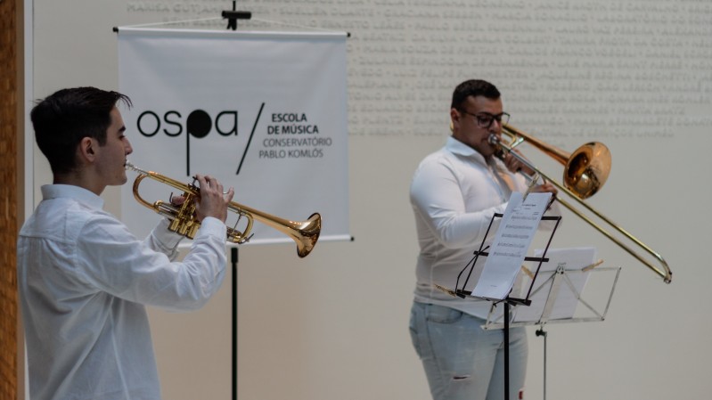 Dois músicos se apresentam em um ambiente interno, vestidos com camisas brancas. À esquerda, um jovem toca trompete dourado, enquanto à direita, um homem toca trombone de vara. Ambos estão concentrados em sua performance, com partituras posicionadas em estantes à frente deles. Ao fundo, há um banner com o logotipo da OSPA (Orquestra Sinfônica de Porto Alegre) e os dizeres “Escola de Música / Conservatório Pablo Komlós”