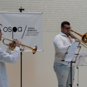 Dois músicos se apresentam em um ambiente interno, vestidos com camisas brancas. À esquerda, um jovem toca trompete dourado, enquanto à direita, um homem toca trombone de vara. Ambos estão concentrados em sua performance, com partituras posicionadas em estantes à frente deles. Ao fundo, há um banner com o logotipo da OSPA (Orquestra Sinfônica de Porto Alegre) e os dizeres “Escola de Música / Conservatório Pablo Komlós”