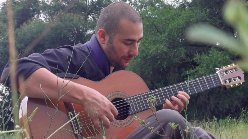 A imagem mostra o instrumentista André Gonçalves, homem sentado ao ar livre, tocando um violão clássico. Ele está concentrado, olhando para as cordas do instrumento enquanto toca. O homem tem cabelo curto raspado e barba rala. Está vestido com uma camisa escura com lenço azul. Ao fundo, há árvores verdes e vegetação, sugerindo que ele está em um campo ou parque. A atmosfera da imagem é tranquila e introspectiva.