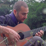 A imagem mostra o instrumentista André Gonçalves, homem sentado ao ar livre, tocando um violão clássico. Ele está concentrado, olhando para as cordas do instrumento enquanto toca. O homem tem cabelo curto raspado e barba rala. Está vestido com uma camisa escura com lenço azul. Ao fundo, há árvores verdes e vegetação, sugerindo que ele está em um campo ou parque. A atmosfera da imagem é tranquila e introspectiva.
