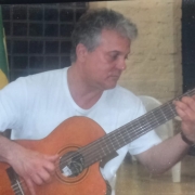 Foto do violonista Onésimo Duarte. Ele é um homem de cabelos grisalhos e camiseta branca e está tocando um violão de forma concentrada. Ele está em um ambiente interno, com cadeiras de plástico ao fundo e bandeiras (uma delas verde e amarela) visíveis parcialmente. 