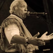 Foto de Marco Aurélio Vasconcellos, de cabelos grisalhos e expressão intensa, cantando em um microfone. Ele veste uma camisa clara e um poncho com franjas, característico da indumentária gaúcha. A foto é em preto e branco, durante uma apresentação ao vivo, com um fundo escuro e equipamentos de som visíveis.

