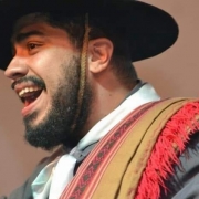 Foto do cantor Jader Leal, segurando um microfone. Ele usa uma vestimenta tradicional gaúcha, com lenço no pescoço e um poncho com detalhes vermelhos e pretos. O chapéu de abas largas completa o traje típico. A imagem transmite energia e paixão pela música.