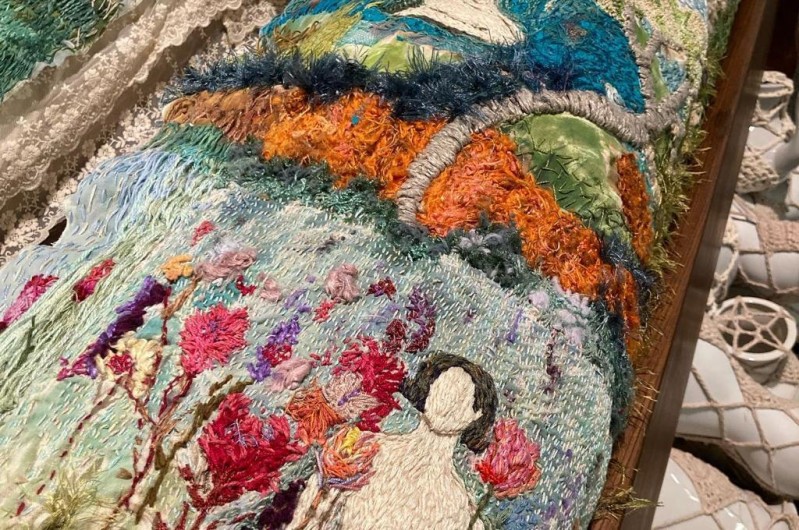 Imagem de um bordado artístico feito com diversas texturas e fios coloridos. A cena central mostra uma figura feminina nua, sentada em uma cadeira de encosto alto com assento bordô. Ela se inclina para a frente, com as mãos e os pés tocando um fio de água azul que flui por uma pequena queda.