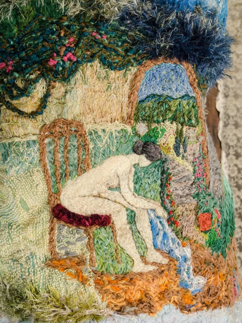 Imagem de um bordado artístico feito com diversas texturas e fios coloridos. A cena central mostra uma figura feminina nua, sentada em uma cadeira de encosto alto com assento bordô. Ela se inclina para a frente, com as mãos e os pés tocando um fio de água azul que flui por uma pequena queda.