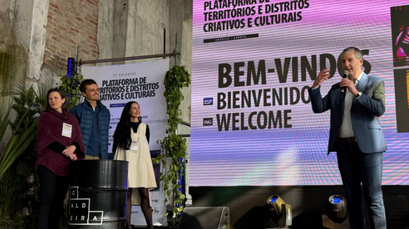Quatro pessoas estão em um palco, em frente a uma tela digital que exibe “BEM-VINDOS / BIENVENIDOS”. À esquerda, uma mulher de suéter vinho sorri; ao lado dela, uma mulher de vestido branco e blusa preta olha atentamente; um homem de terno azul está ao lado, e outro homem de jaqueta azul e microfone nas mãos fala ao público. Ao fundo, um banner vertical diz “Plataforma de Territórios e Distritos Criativos e Culturais”.