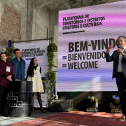 Quatro pessoas estão em um palco, em frente a uma tela digital que exibe “BEM-VINDOS / BIENVENIDOS”. À esquerda, uma mulher de suéter vinho sorri; ao lado dela, uma mulher de vestido branco e blusa preta olha atentamente; um homem de terno azul está ao lado, e outro homem de jaqueta azul e microfone nas mãos fala ao público. Ao fundo, um banner vertical diz “Plataforma de Territórios e Distritos Criativos e Culturais”.

